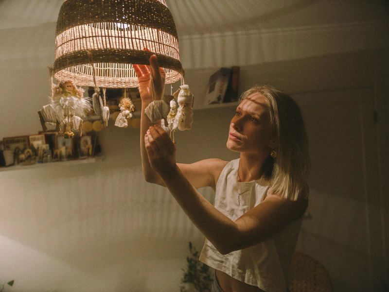Mulher ajustando um móbile sob a luz baixa de uma luminária de palha, em um quarto